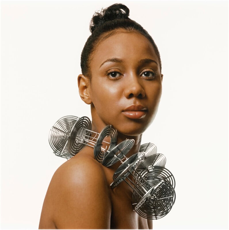 A woman with a neat bun poses against a white background, wearing a large, sculptural necklace made of intertwined metal coils around her neck and shoulders.