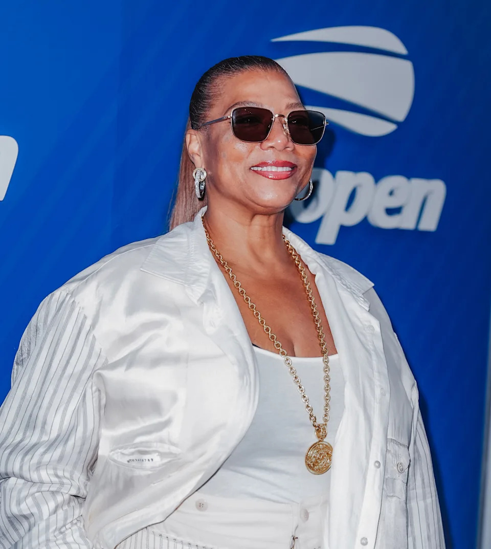 Queen Latifah in stylish light-colored jacket and sunglasses poses at a sports event backdrop