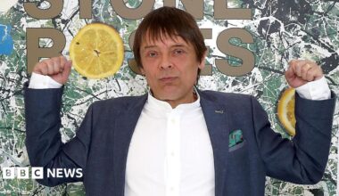 Mani with both arms raise standing in front of a large image of the Stone Roses' debut album artwork
