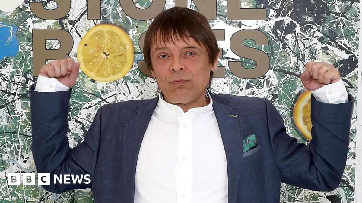 Mani with both arms raise standing in front of a large image of the Stone Roses' debut album artwork