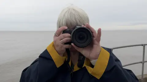 BBC Liz Elmont is pointing her camera lens at the viewer. She has short white hair and is standing in front of the sea, which is grey.