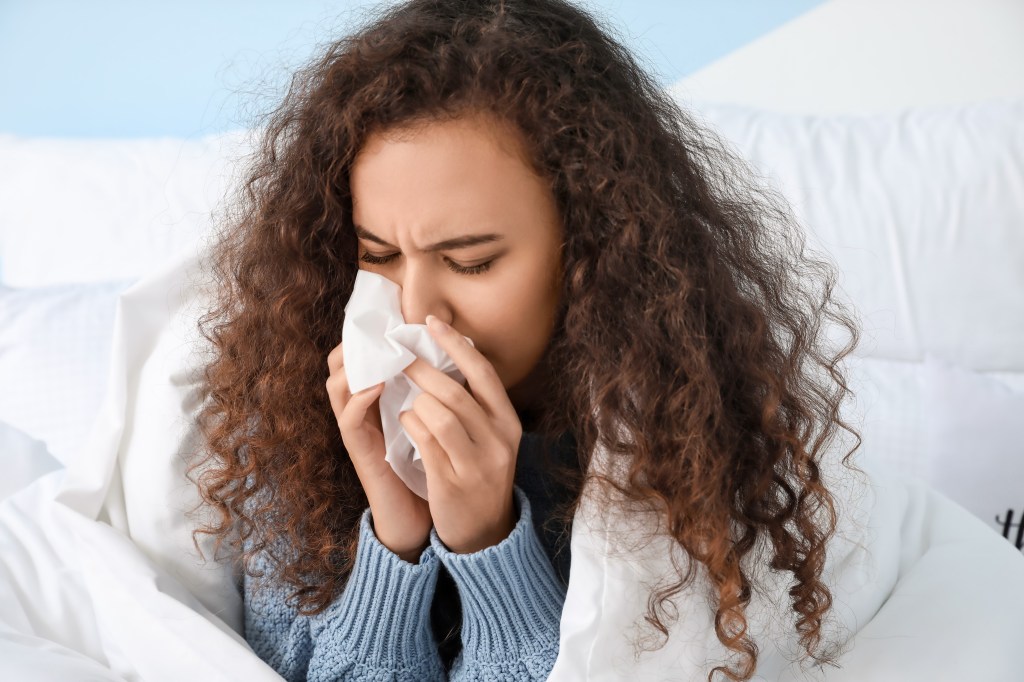 African American woman with curly hair blowing her nose into a tissue in bed, covered in a white blanket.