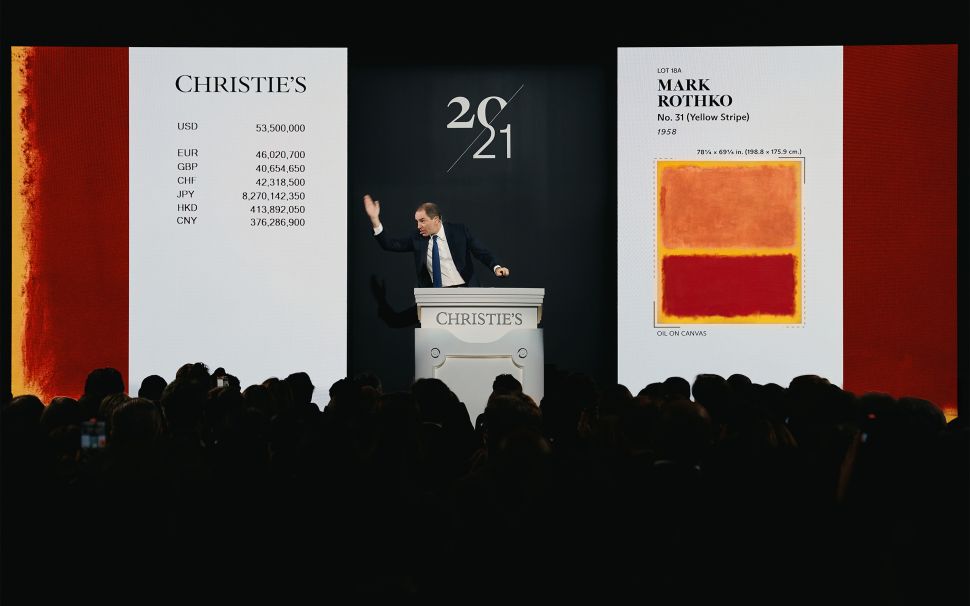 A Christie’s auctioneer gestures from the podium as Mark Rothko’s No. 31 (Yellow Stripe) and its multimillion-dollar currency conversions are displayed on large screens before a packed salesroom.
