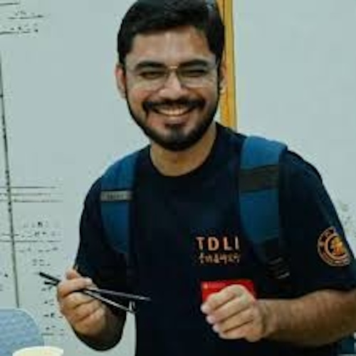 Smiling young man with dark hair and short beard, wearing a TDLI t-shirt and holding chopsticks.
