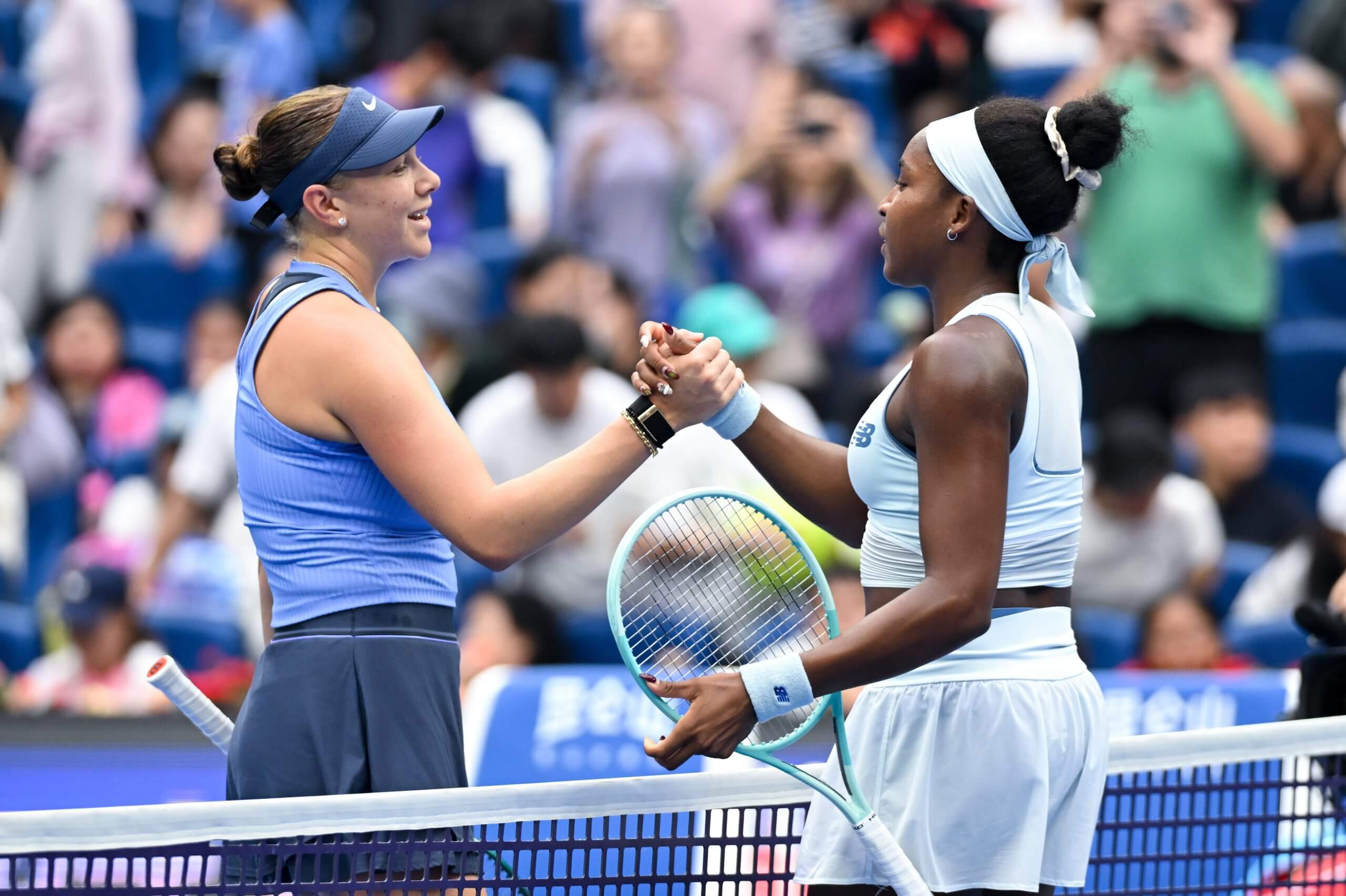 Amanda Anisimova (left) wearing a blue visor and outfit and Coco Gauff (right) wearing a white headband and outfit shake hands across a tennis net.