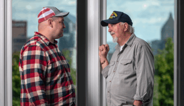 Two male Veterans, one in a red plaid shirt and cap and the other in a gray shirt and navy cap, face each other by a window, appearing deep in conversation. Both are Peer Specialists.