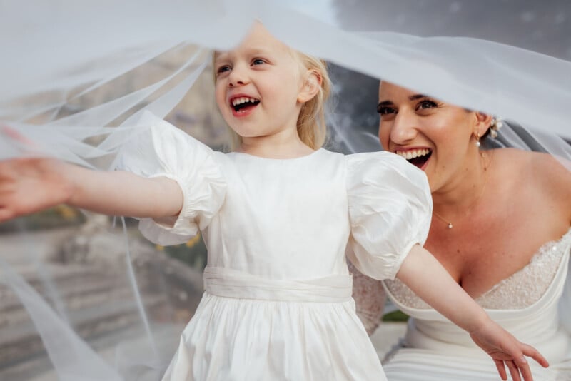 A joyful bride and a smiling young girl in white dresses playfully hold up a flowing bridal veil outdoors, both looking happy and excited.