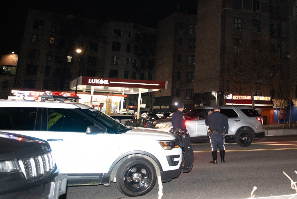 Police investigate an incident on a street at night, with two police officers standing by a silver SUV and a police car in the foreground, next to a Lukoil gas station.