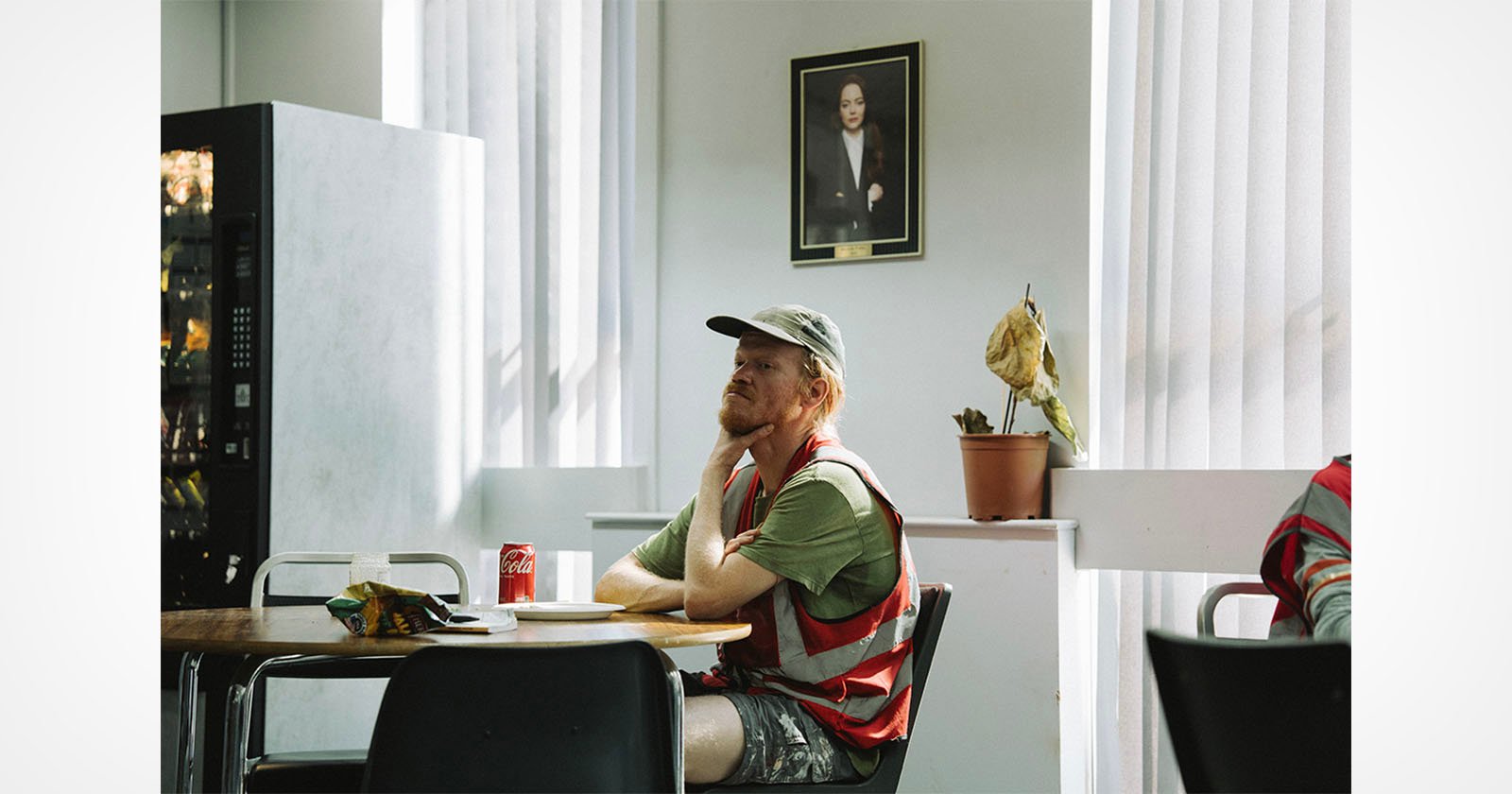 A man in a reflective vest sits at a table with snacks and a can of Coke, appearing thoughtful. Sunlight filters through vertical blinds. A vending machine, potted plant, and a framed portrait are in the background.