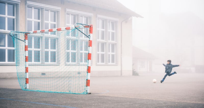 A child kicks a soccer ball toward a goal on an empty schoolyard, with fog partially obscuring the building in the background.