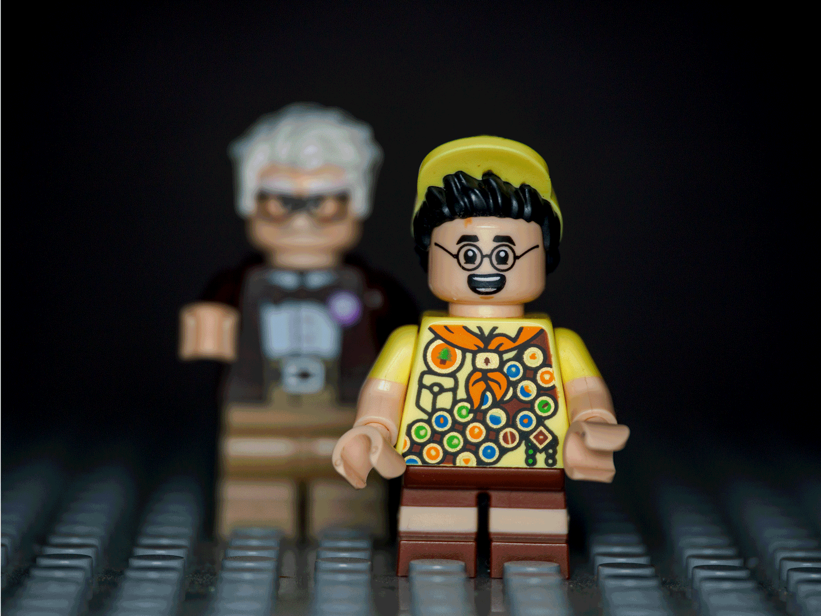 Two LEGO minifigures stand on a gray surface; the front figure wears glasses, a yellow hat, and a colorful shirt, while the blurred figure in the background has white hair and dark clothing.