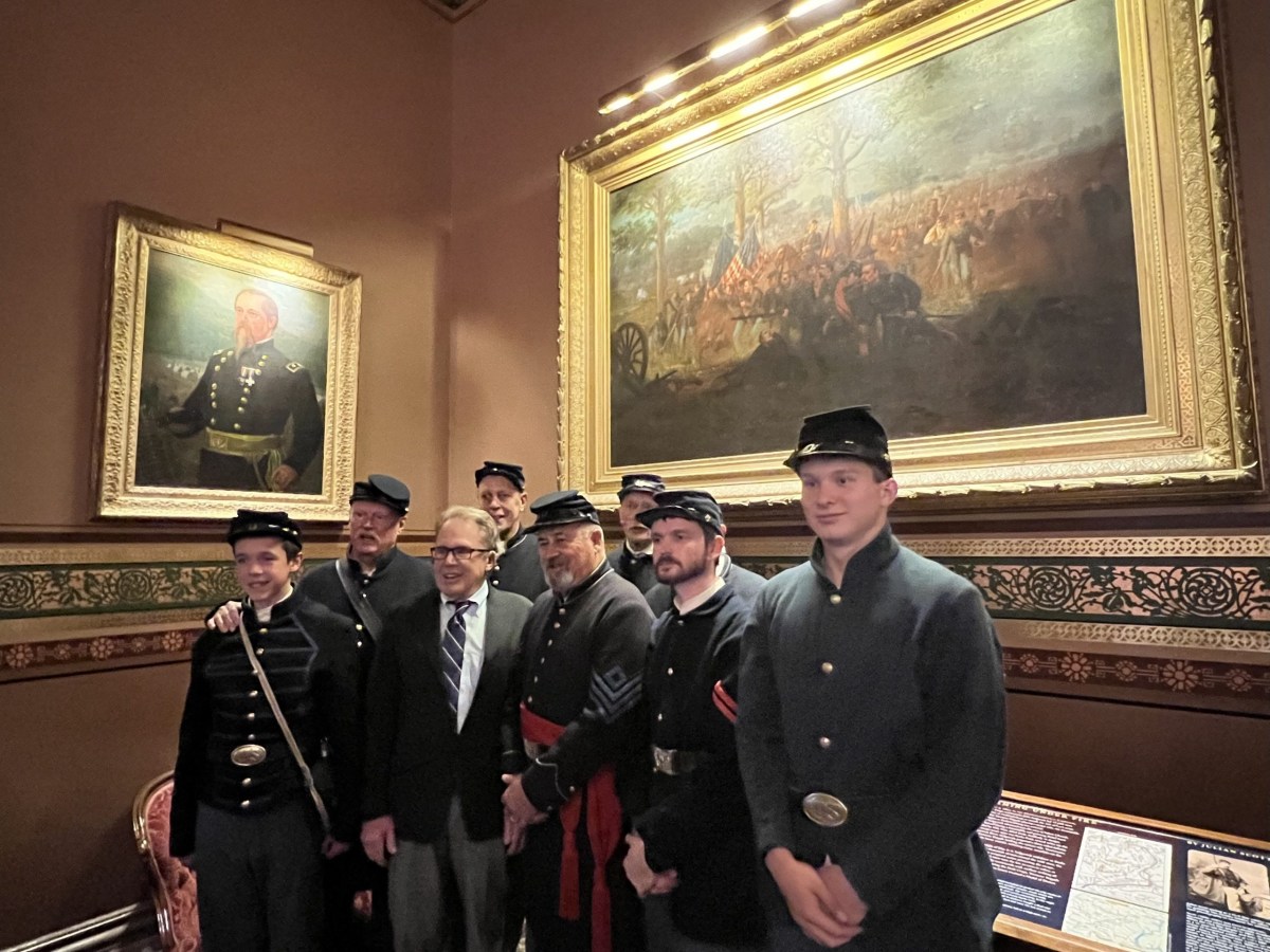 A group of men, some in Civil War-era uniforms, pose for a photo indoors beneath two large framed paintings in a historic room.