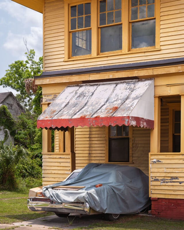 A vintage yellow wooden house with peeling paint has a weathered awning over a classic car parked in front, partially covered by a blue tarp. Lush greenery and another old house appear in the background.
