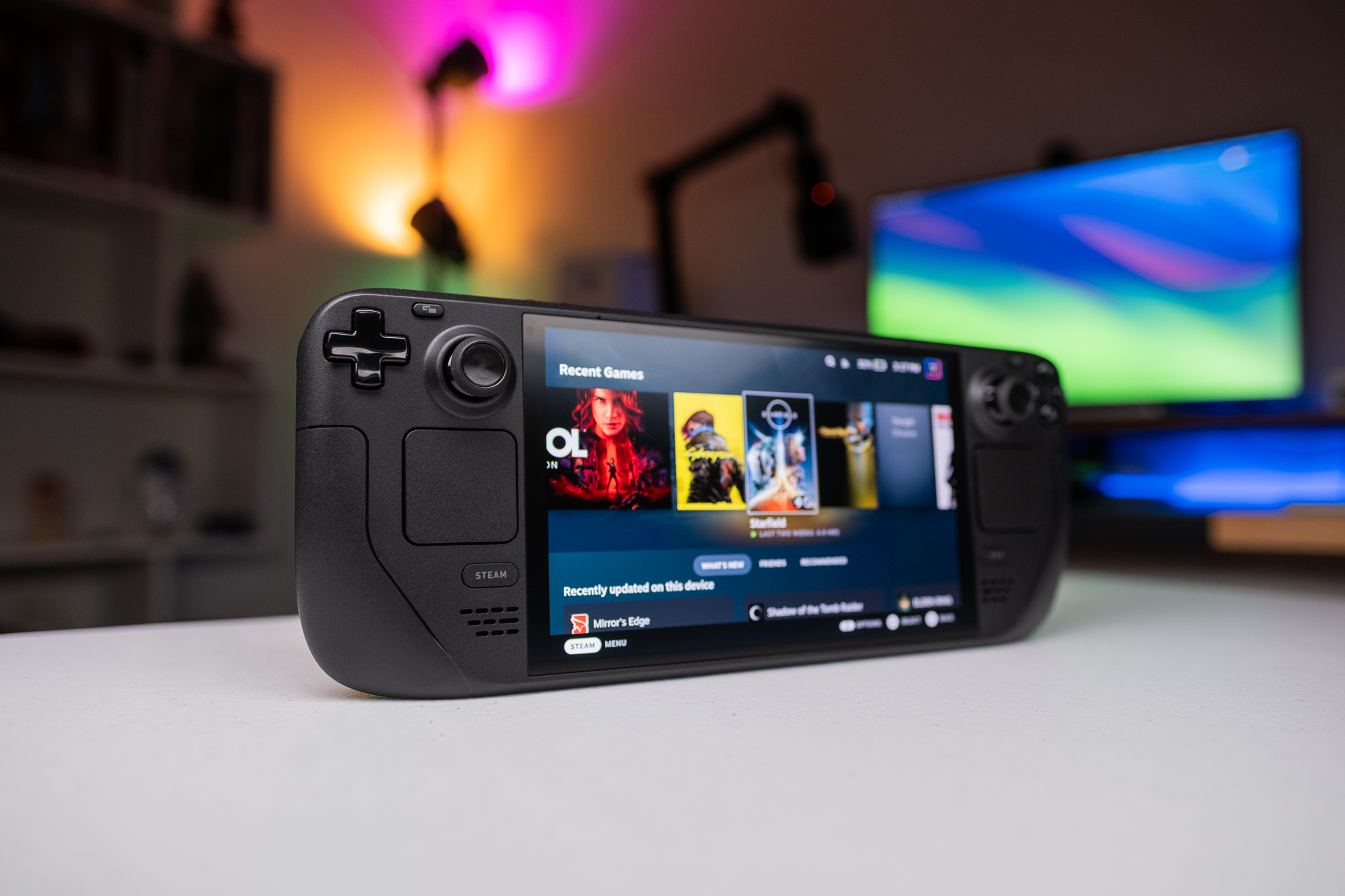The Steam Deck gaming handheld sits on a white table, illuminated by rainbow light, showcasing a library of games on its screen including Control, Cyberpunk and Starfield.