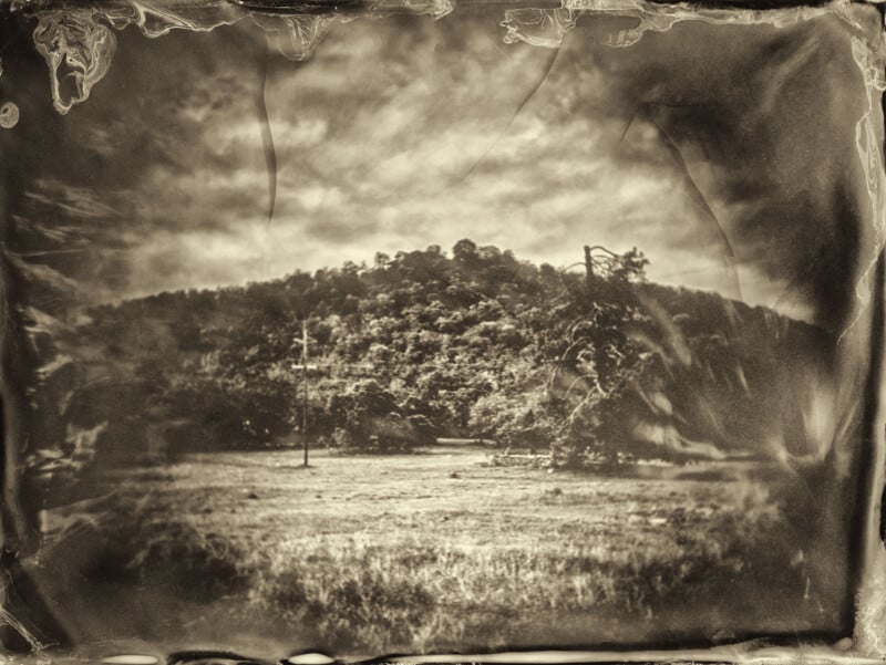 Sepia-toned, vintage-style photograph of a grassy field with a tree and a forested hill in the background, surrounded by a worn, scratched, and textured border. The scene appears aged and atmospheric.