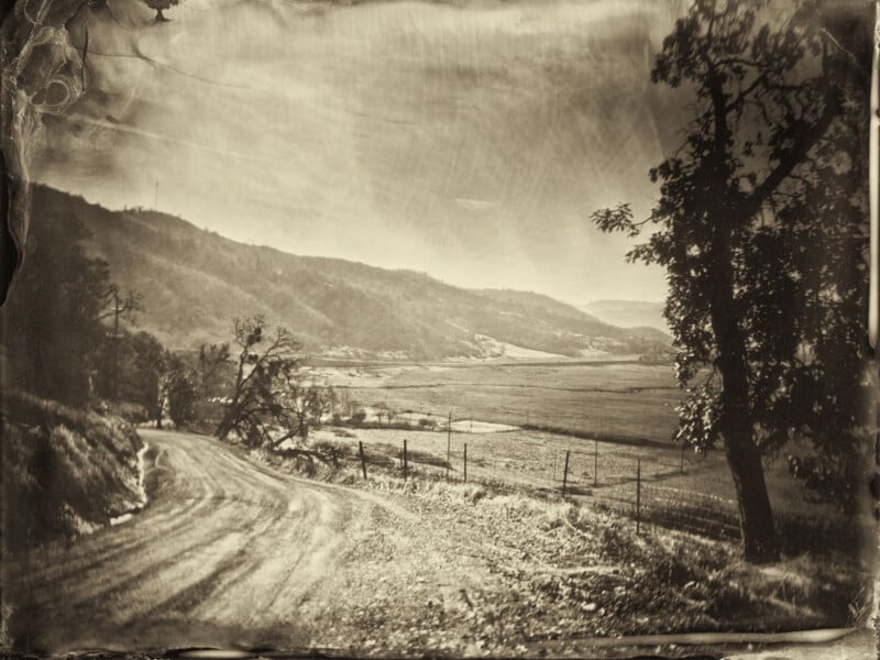 Sepia-toned photograph of a winding dirt road curving through a rural landscape, bordered by hills and fields, with trees framing the scene and distant mountains in the background.