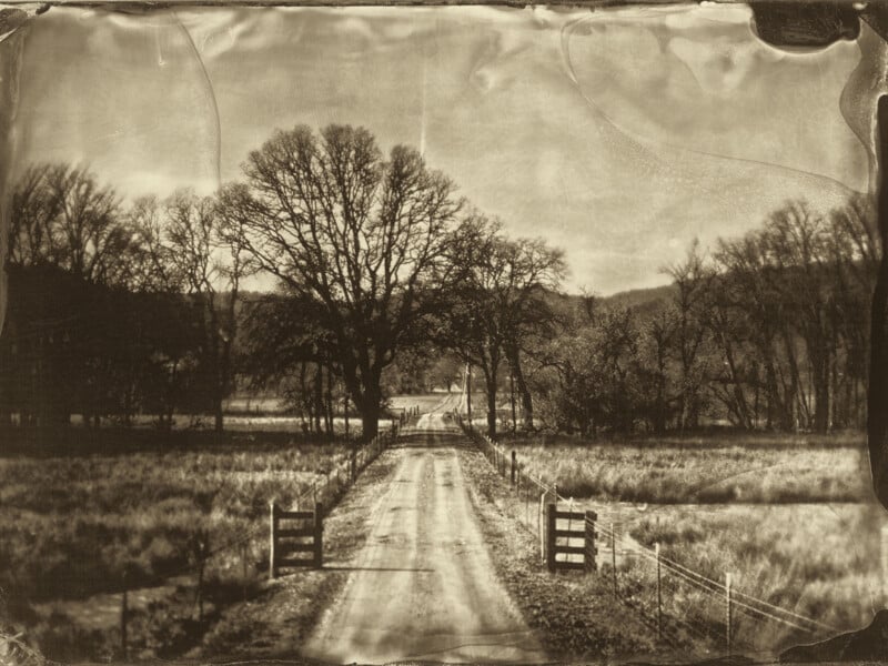 A sepia-toned, vintage-style photo of a rural dirt road flanked by wooden gates and fencing, leading toward distant leafless trees and hills under a cloudy sky.