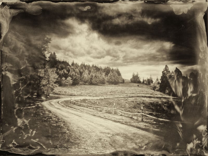 Sepia-toned photograph of a winding dirt road curving through a grassy field with trees on the hillsides under a cloudy sky, resembling an old vintage or wet plate style image.