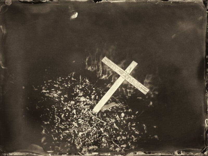 A sepia-toned photo of a wooden cross grave marker in the ground, surrounded by scattered leaves. The cross is slightly tilted and the background is dark and blurred, creating a somber atmosphere.