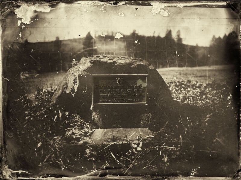 Sepia-toned photograph of a memorial plaque mounted on a large rock, set in a grassy field with trees and hills visible in the blurred background; the image appears old and worn.