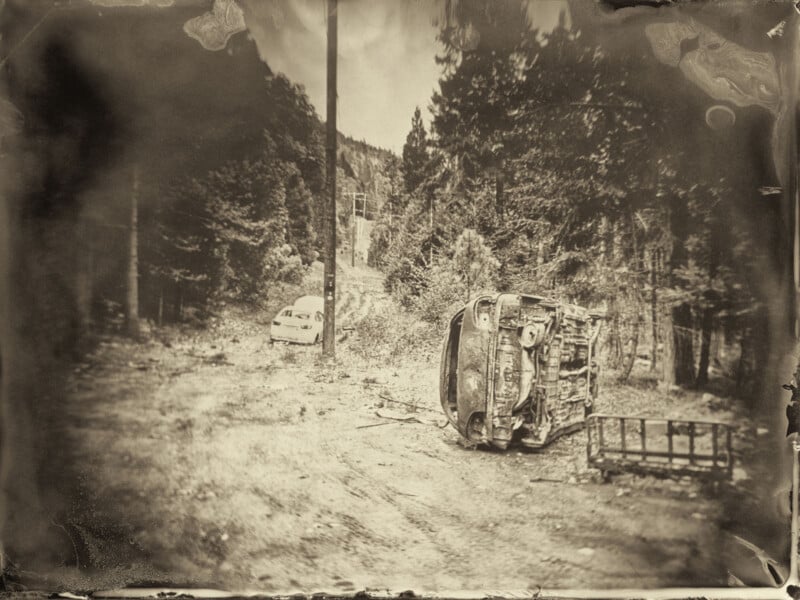 A sepia-toned photo shows a forested dirt road with an overturned car on its side in the foreground and another abandoned car further down the road among tall trees. The image has an old, weathered appearance.