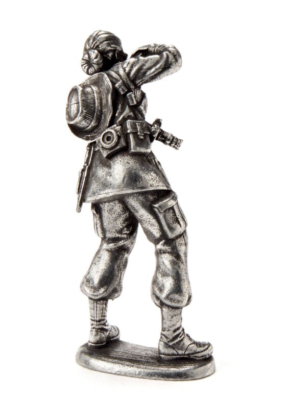 A small metallic figurine of a person wearing a backpack, pants, and boots, standing and holding binoculars to their face, viewed from the back on a plain white background.