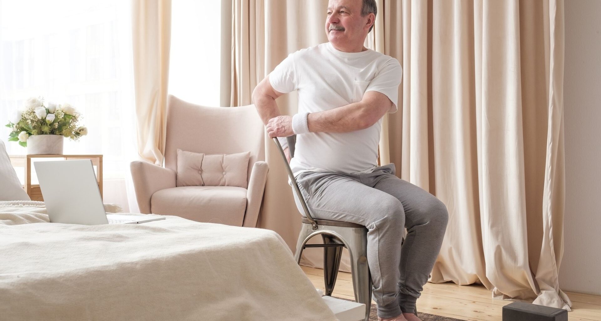 man in a white tshirt and grey joggers sitting in a chair and twisting to one side. he&#039;s in a bedroom setting with a laptop on the bed and a yoga block on the floor.