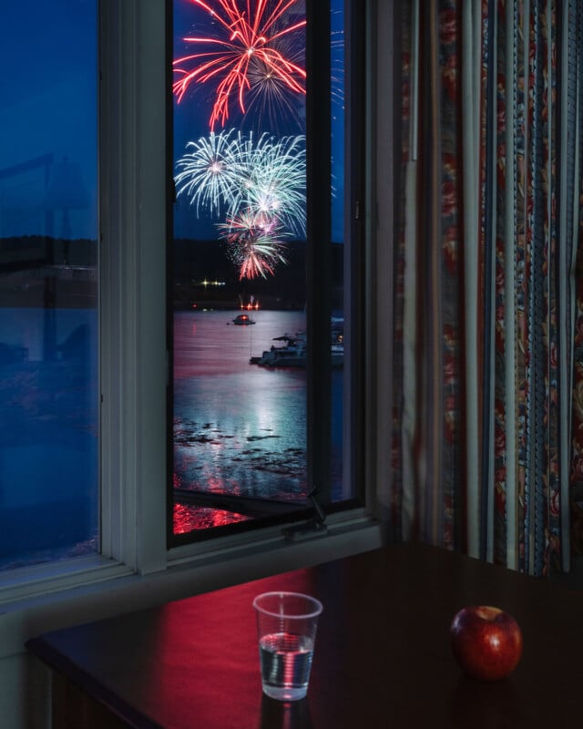 A room with a table holding a clear plastic cup of water and a red apple; through an open window, colorful fireworks light up the night sky over a body of water with boats.