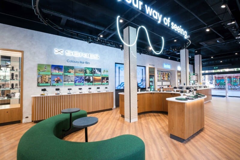 Modern electronics store interior with curved green seating, wooden display counters, and illuminated signs showcasing cameras and lenses. Wall displays feature colorful photos, and large windows let in natural light from outside.