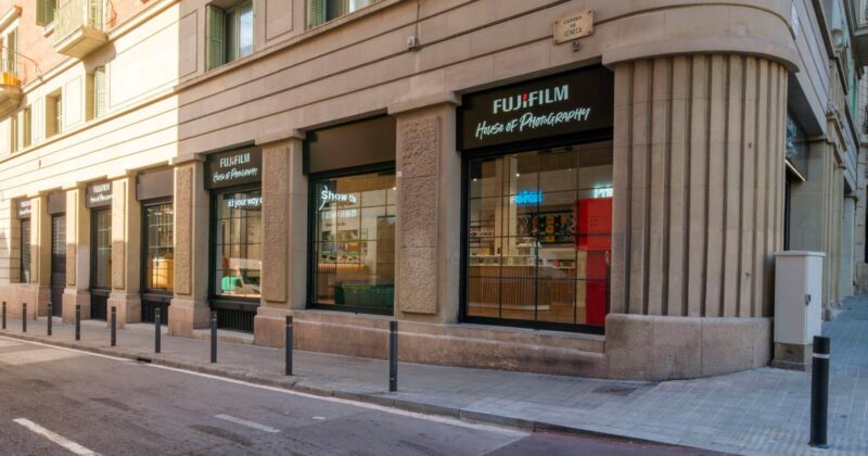 A corner view of a Fujifilm House of Photography store with large windows, black awnings, and a stone facade on a quiet city street. Bollards line the sidewalk in front of the shop.