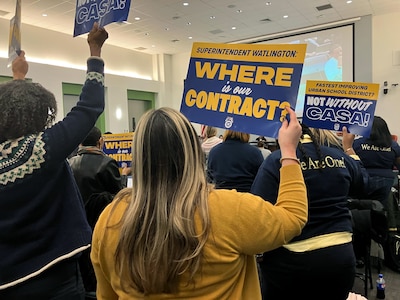 A photograph of a meeting room full of adults, some of them are holding up protest signs.