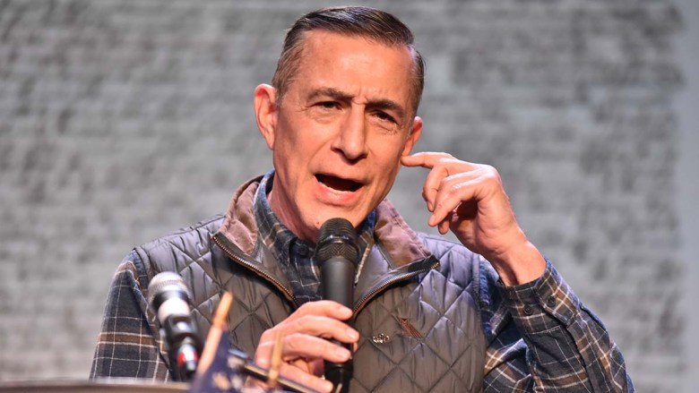 A person wearing a plaid shirt and brown vest speaks into a microphone
