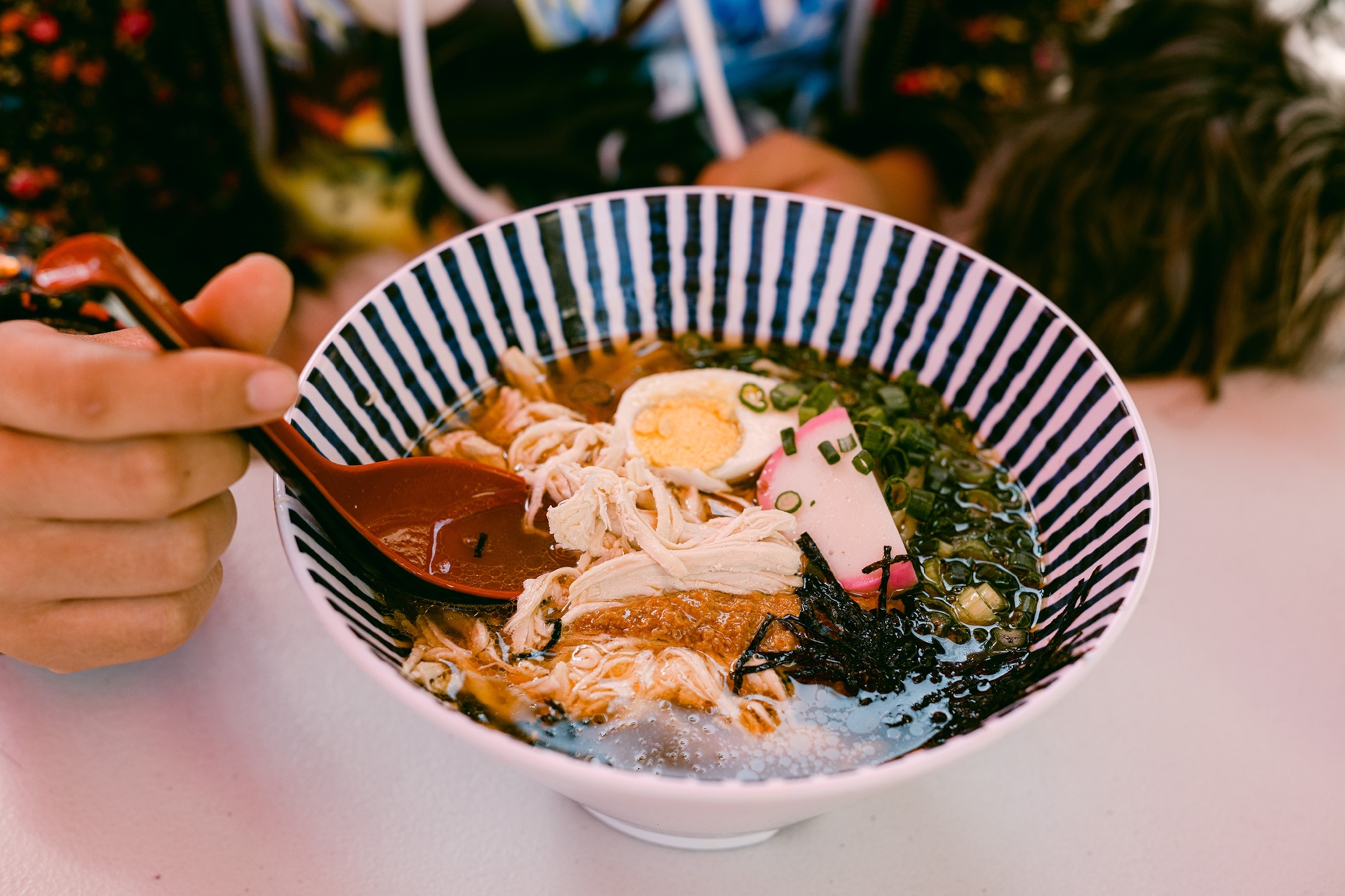 A cloe-up food shot of a funnel-shaped bowl of noodle soup topped with a halved, hard-boiled egg, seaweed strips and pulled chicken.