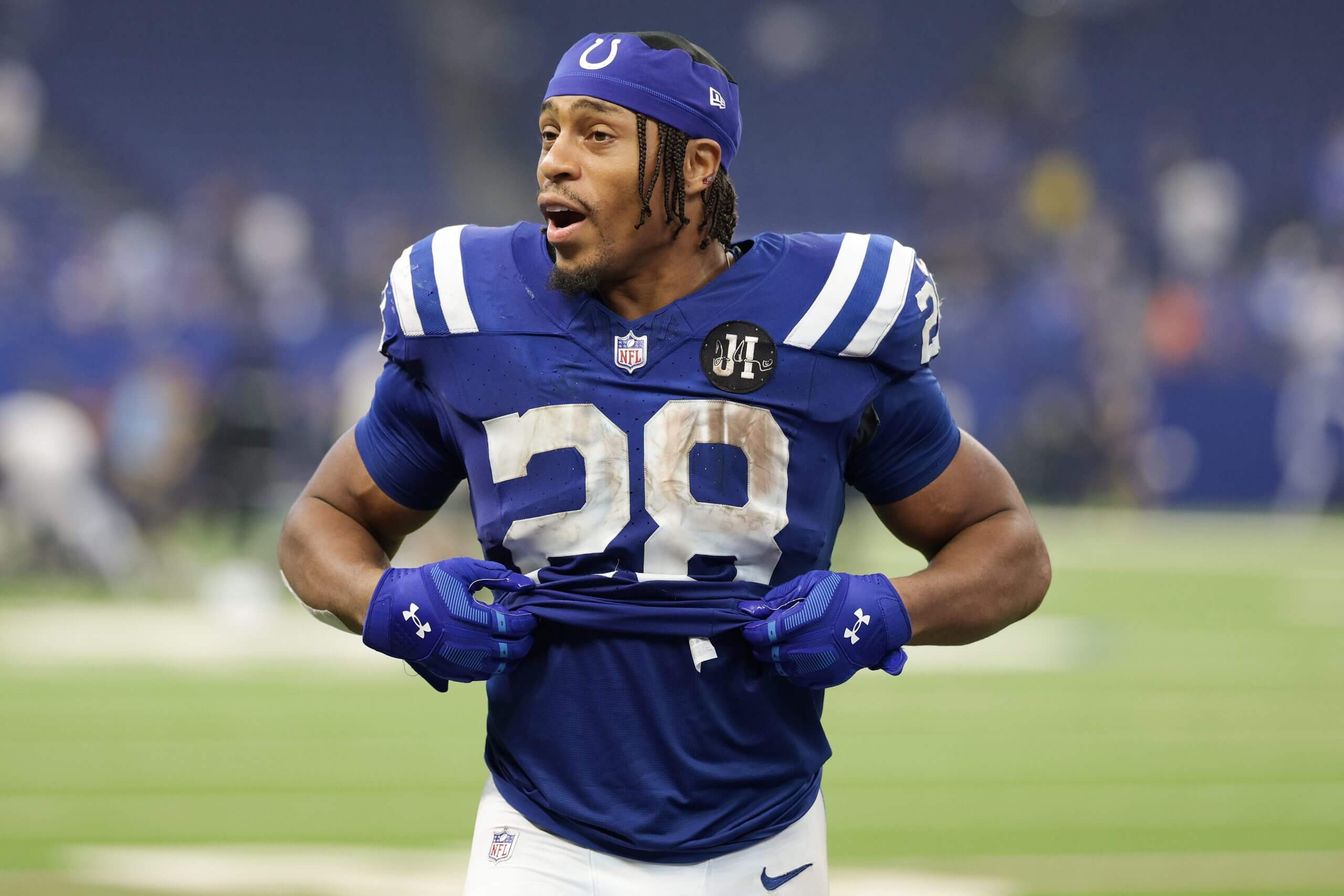 Jonathan Taylor, wearing a blue No. 28 uniform with no helmet, holds his jersey rolled halfway up his stomach, with his mouth open in a questioning expression.
