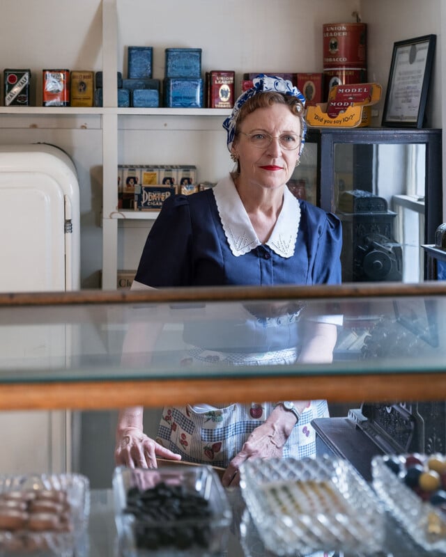 A woman in vintage clothing, including a blue dress, white collar, and patterned apron, stands behind the counter of an old-fashioned shop with glass display trays and shelves of retro products behind her.