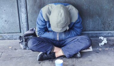 A person wearing a hooded jacket sits on the pavement with their head down and arms crossed over their knees. A backpack and a paper cup are nearby, and the background is a worn, gray wall.