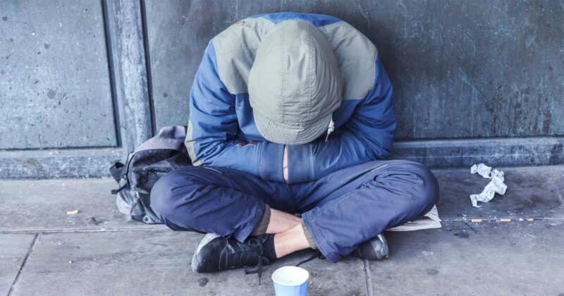 A person wearing a hooded jacket sits on the pavement with their head down and arms crossed over their knees. A backpack and a paper cup are nearby, and the background is a worn, gray wall.