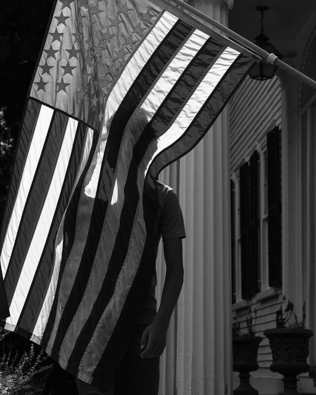 A person stands behind a large American flag, which partially obscures their figure, in front of a building with white columns and shuttered windows. The image is in black and white.