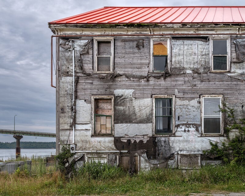 Weathered, abandoned two-story building with peeling paint, broken and boarded-up windows, overgrown grass, and a faded red metal roof, situated near a river with a bridge in the background under a cloudy sky.