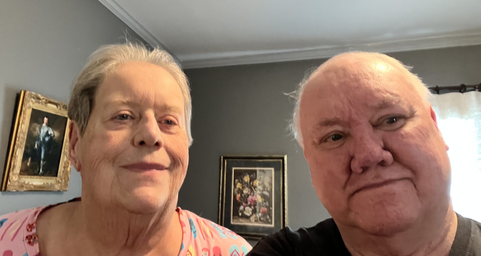 A senior man and woman seated in a room with several framed images behind them