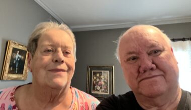 A senior man and woman seated in a room with several framed images behind them
