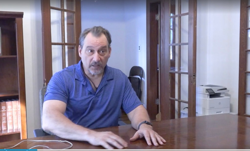 Robert Warnicke, vice president of the Phoenix Historic Neighborhood Coalition, in his law office in La Hacienda, one of 36 neighborhoods effected by a housing plan unanimously approved by the city council. He sits at his table with both hands pressed on top. A doorway is over his left shoulder.
