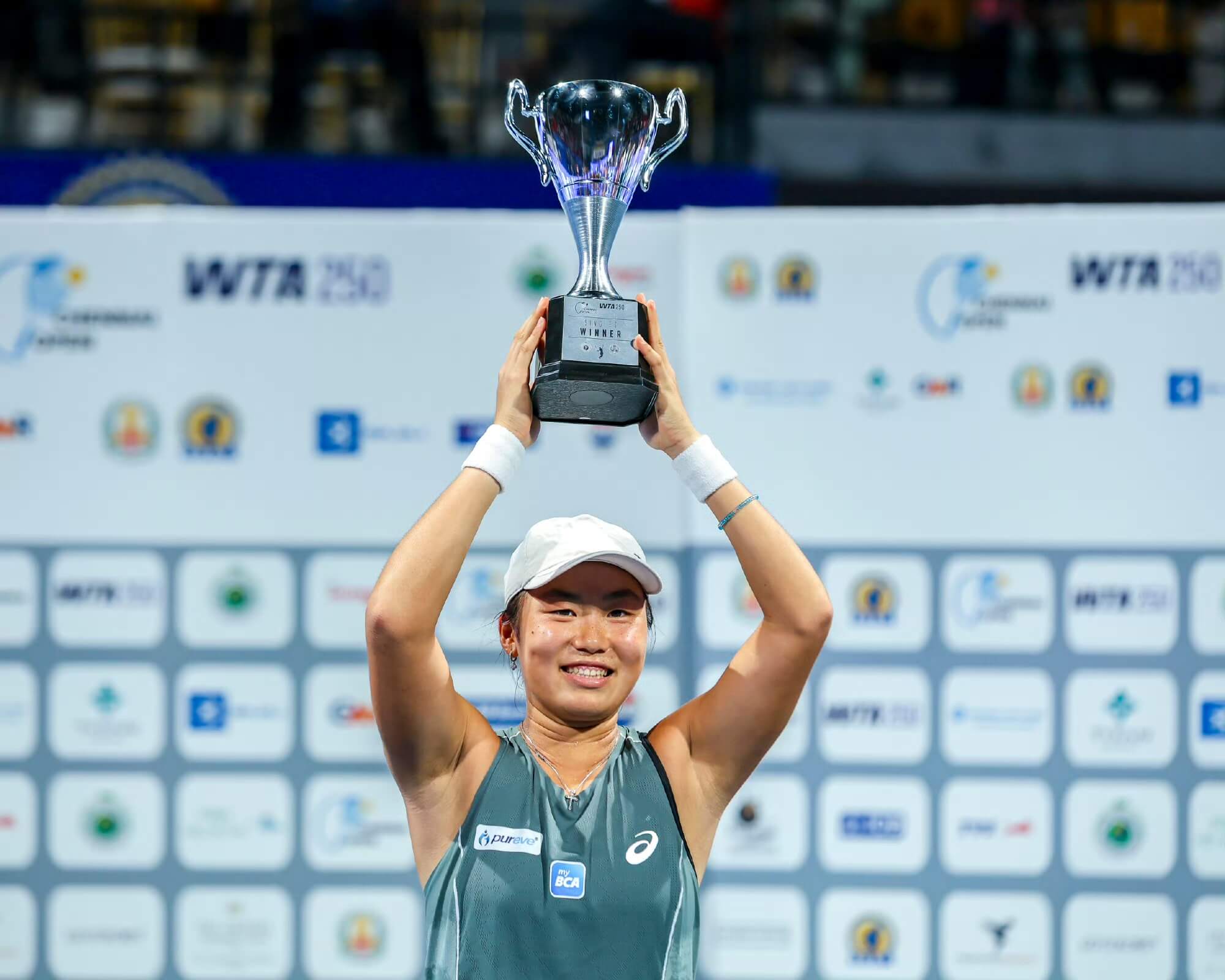 Janice Tjen wears a turquoise tennis outfit and white cap as she holds a silver trophy above her head.