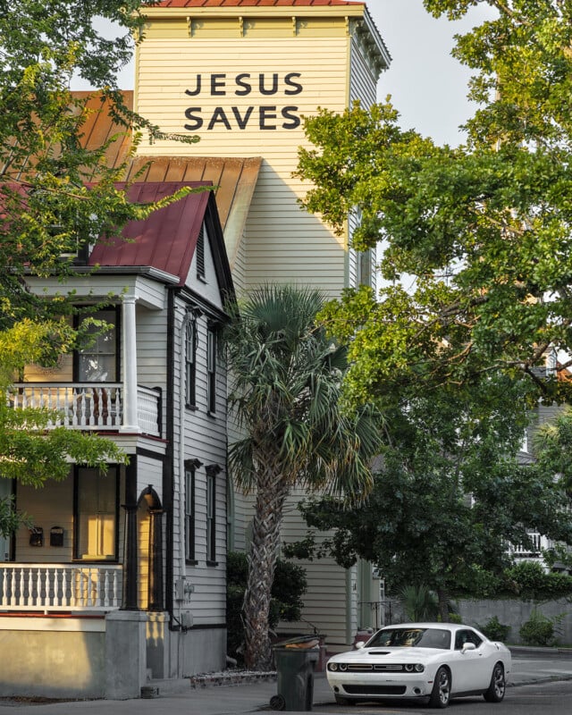 A white car is parked in front of a house with a porch and palm tree. Behind it, a tall building displays a large sign reading “JESUS SAVES.” Green trees frame the scene.