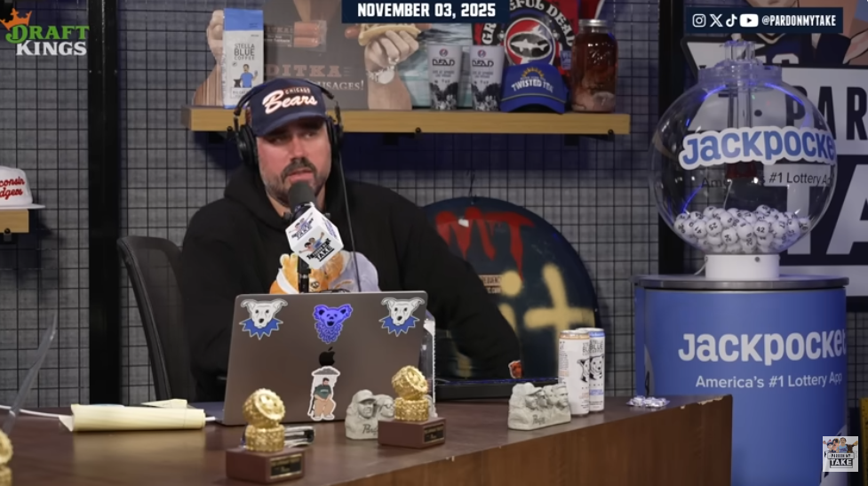 Katz sitting at a desk with a microphone and laptop, next to a lottery machine.