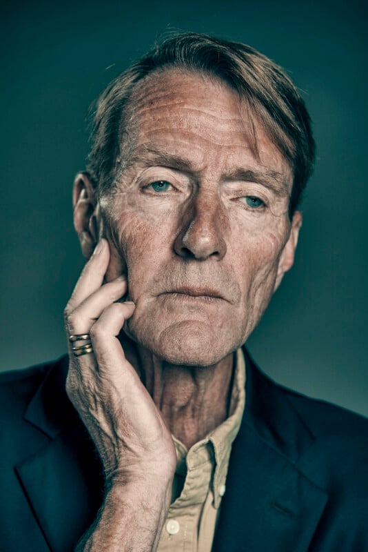 A mature man with light skin and short, light brown hair gazes thoughtfully, resting his hand on his cheek. He wears a dark blazer, light shirt, and a gold ring, set against a plain, dark background.
