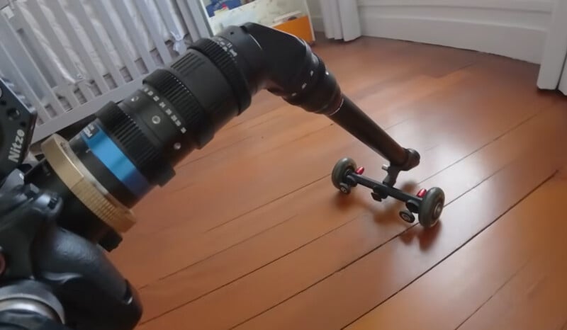 A camera with a long lens is mounted on an adjustable arm attached to a small wheeled dolly, positioned on a wooden floor near a crib and white walls.