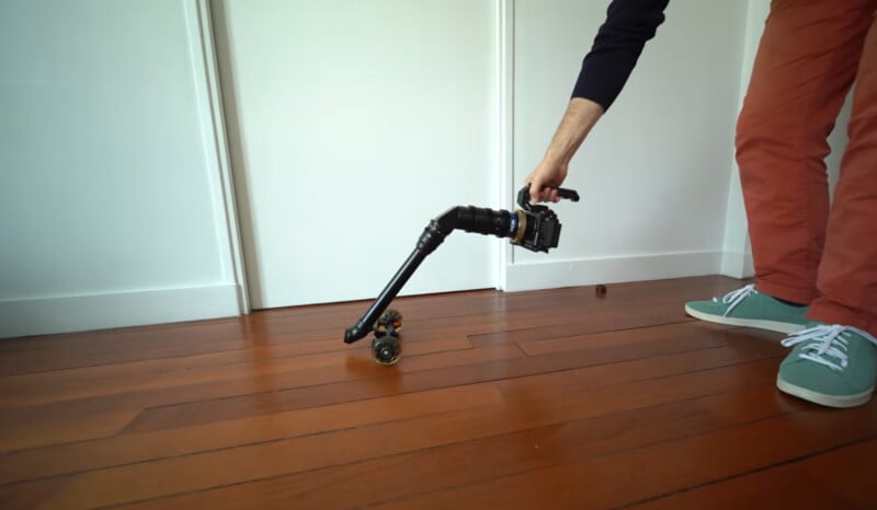 A person wearing red pants and green sneakers holds a camera mounted on a small dolly with an angled arm, positioned low above a wooden floor in front of a white door.