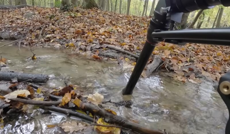 A tripod stands in a shallow forest stream, surrounded by fallen autumn leaves and branches, with trees in the background.