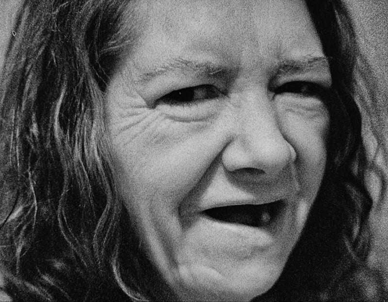 A close-up black and white photo of a middle-aged woman with long wavy hair, wrinkled skin, and a gap in her teeth. She appears to be reacting with surprise or discomfort.